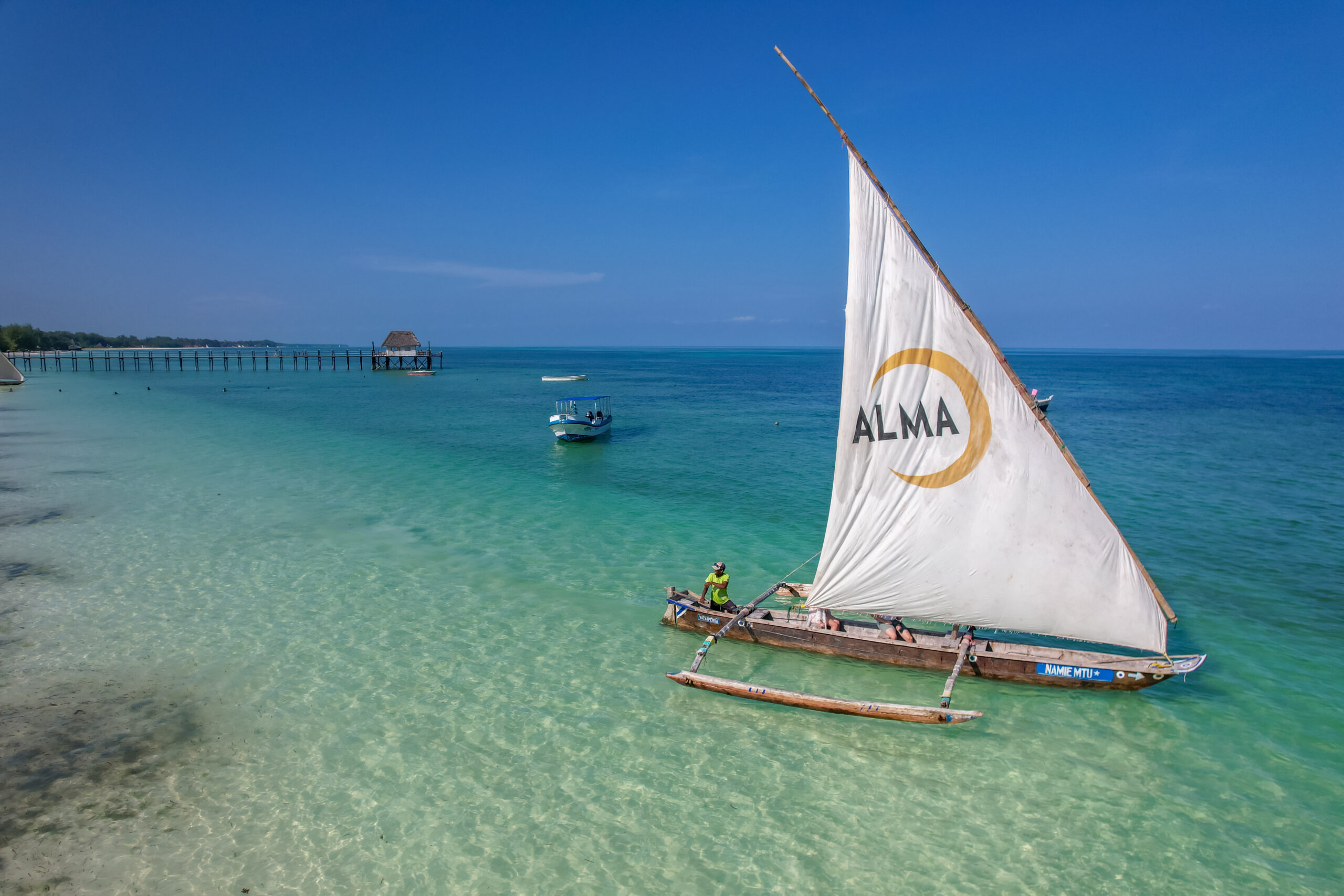 Traditional sailboat with 'ALMA' logo on the sail, floating on clear blue waters near Alma Boutique Hotel, with a wooden pier and distant shoreline in the background.